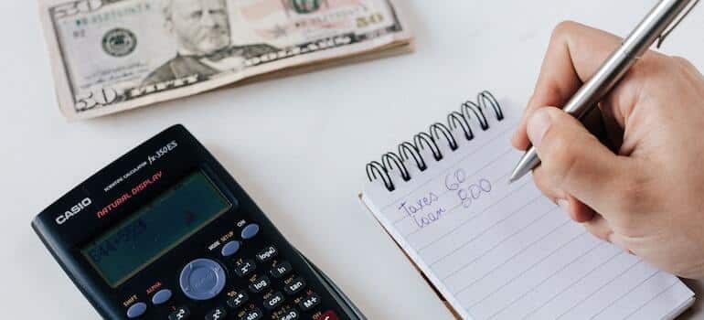 A person writing numbers in a notepad with a calculator and a stack of money besides the notepad
